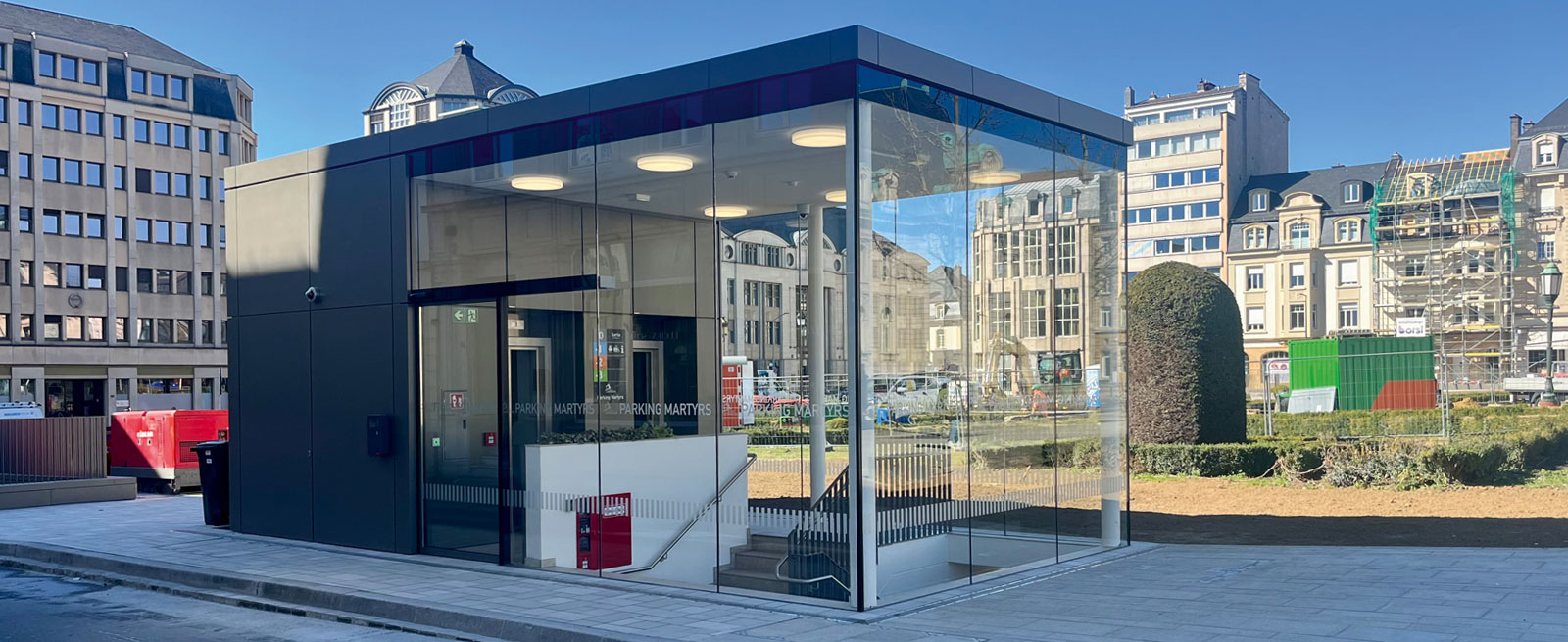 Aussenansicht mit Glassfassade sowie Blechverkleidung vom Parkhauszugang. Im Hintergrund begrünter Platz sowie verschiedene Gebäude.